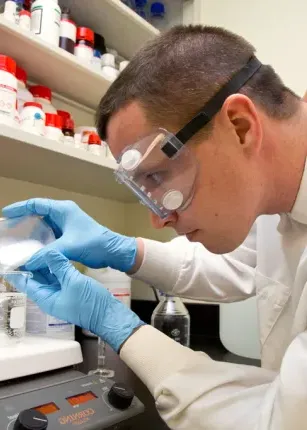 A C O M student pours a substance into a liquid-filled beaker on a scale